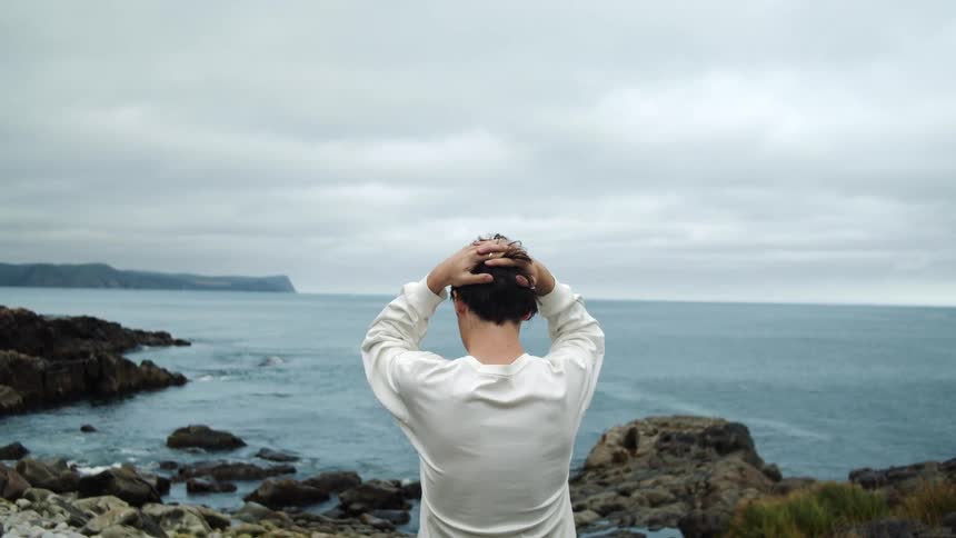 A man stands with hands on his head looking out at the ocean below on a cloudy day