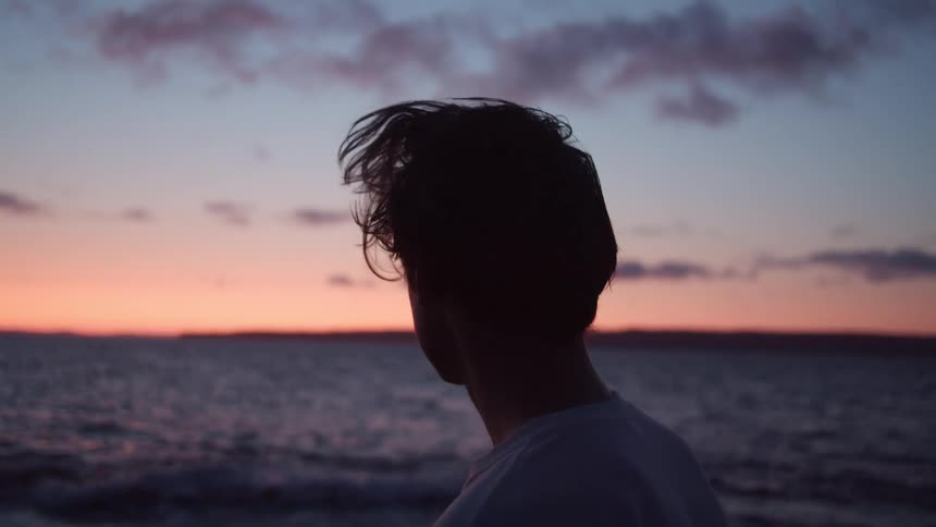 A young man looks at the ocean at sunset, his hair blowing in the wind.
