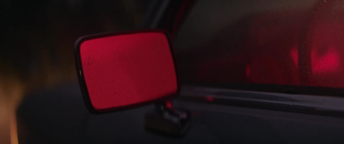 A car side mirror reflects a red light in the rain