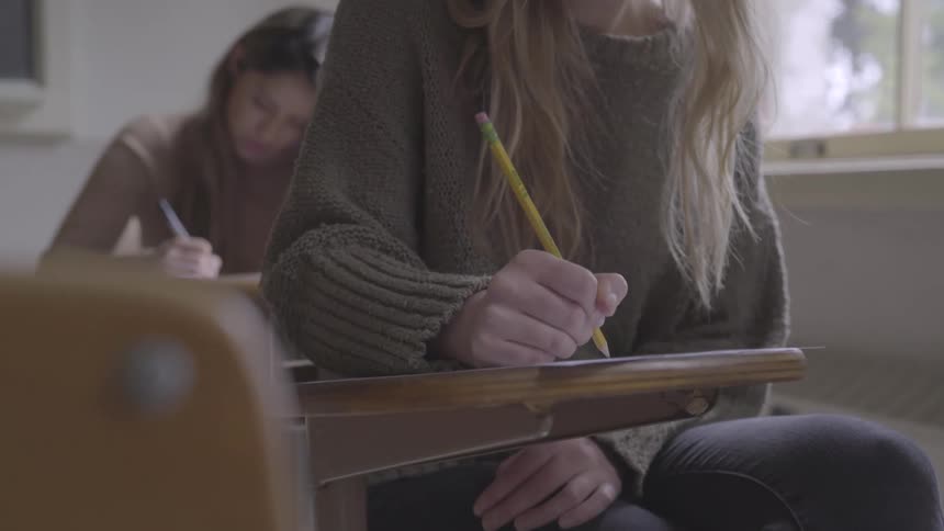 A teenager girl sits at a school desk using a pencil and writing on a piece of paper