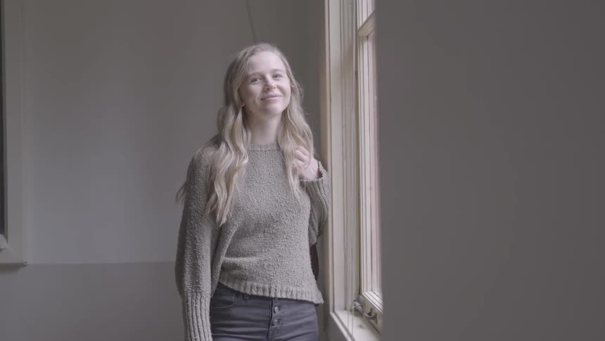Blonde woman with a grey pullover looking out the window turns her gaze to the camera