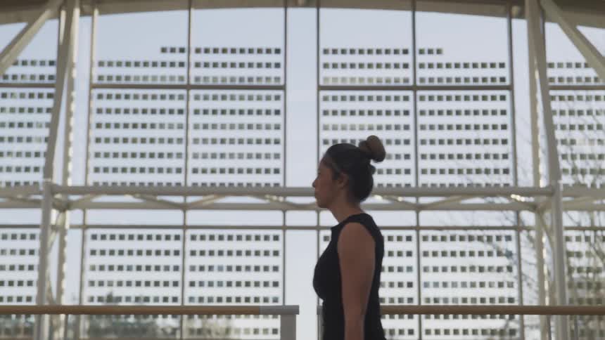 Woman wearing black sleeveless top walks past window showing opposing building and trees