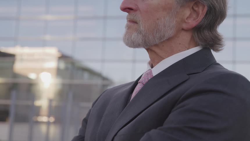 Side view of a businessman with a beard and a suit stands arms crossed in front of an building