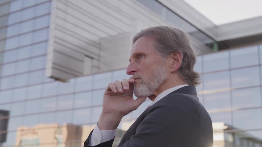 Side view of a businessman with a beard and a suit holding his chin in front of a building