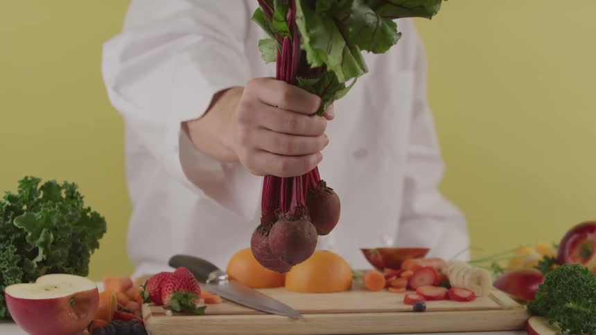 A person with a white shirt holding vegetables over a table with a cutting board a knife fruit and vegetables