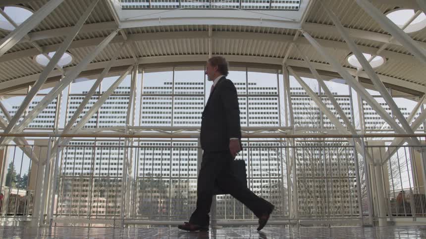 A man in a black suit carries a briefcase as he walks across the top floor of an office building past a large window