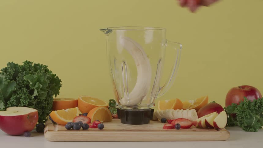 A person places fruit in a blender placed atop a wooden cutting board surrounded by fruit and vegetables