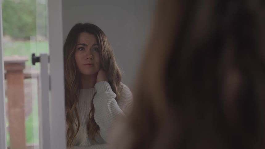 Woman with long brown hair and a grey pullover holds her neck and looks in the mirror