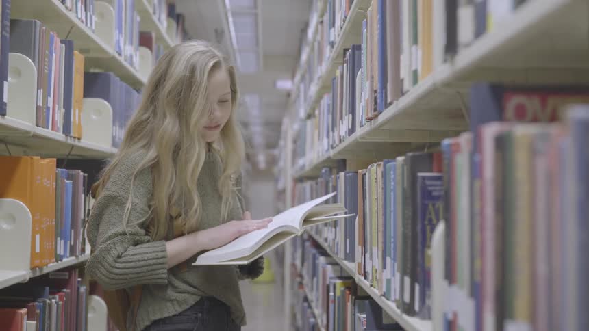 Blonde girl with a grey pullover and a brown backpack looks through a book in the library
