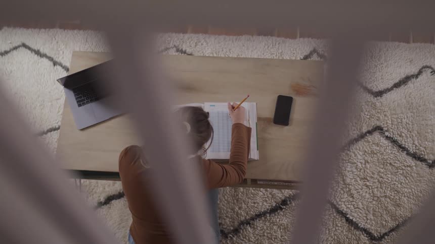 A young woman sits at a desk where she focuses on writing in a workbook