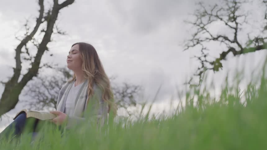 A teenage girl sits in the grass holding a book open and closes her eyes