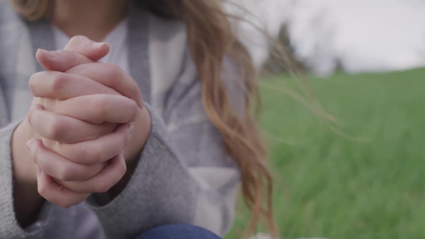 Woman with brown hair and a grey sweater sits on green grass with hands clasped