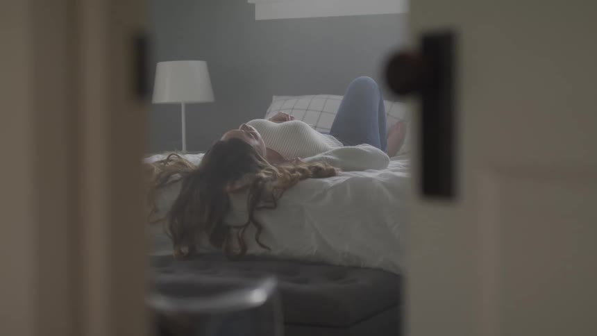 Woman with wavy brown hair a white sweater and jeans lies on bed in bedroom
