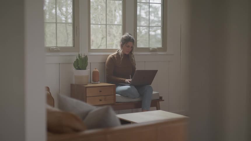 Woman with brown hair a sweater and jeans sits in an armchair and uses a laptop in the living room at home