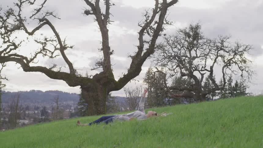 Woman laying down in the grass of a hill near to the trees raises her hand to the sky
