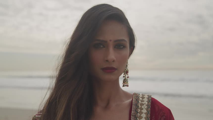 Hindi woman with a bindi on her forehead stands on the sand beach at sunset next to the ocean
