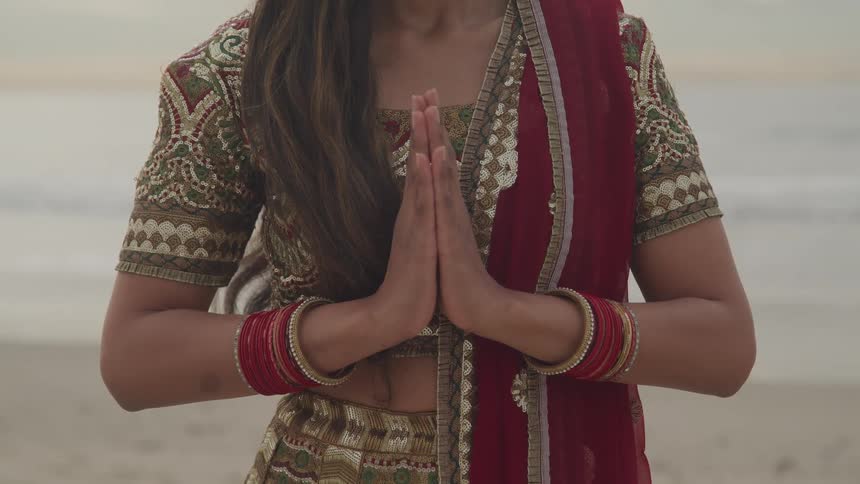 An indian woman in a saree presses her hands together in a namaste greeting while standing on a beach during the day