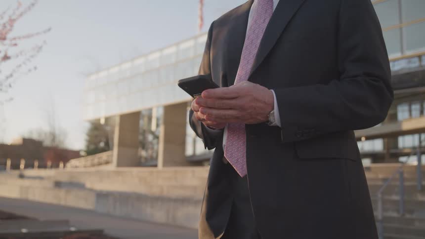 Businessman wearing a suit with a pink tie uses a smartphone outside of the office building
