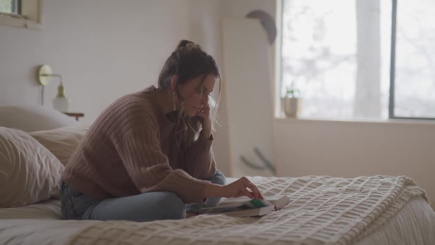 Woman with a pullover and jeans sits on a bed and reads a book in bedroom