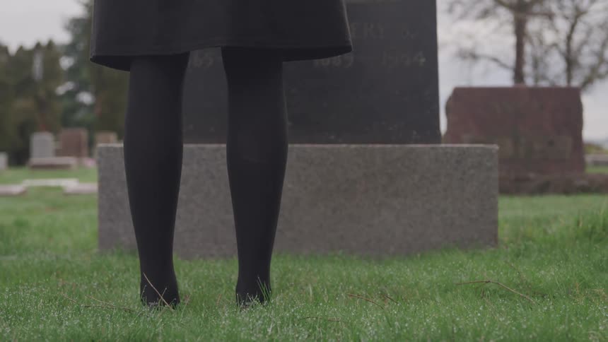 Woman stands in front of grave stone in grass covered conifer bordered cemetery