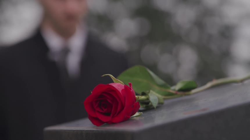 A tombstone with a rose on it with a man with a suit and tie standing in the background