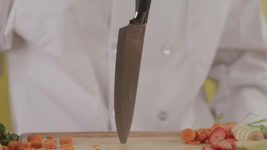 A chef drives a knife point first into a wooden cutting board covered in sliced fruits and vegetables