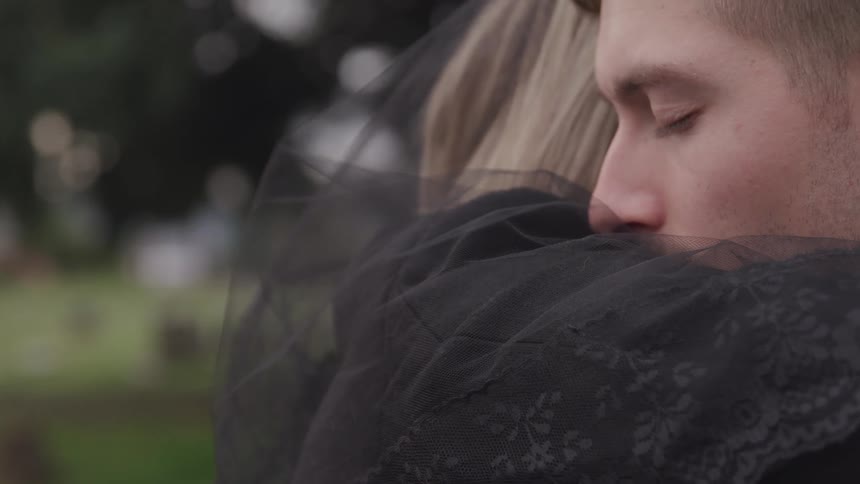 Woman wearing mourning black hugs man in cemetery