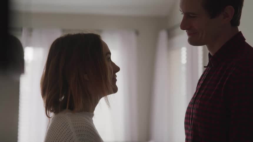 A woman slows raises her head to look into the smiling face of a man as they two of them stand in the living room of a house