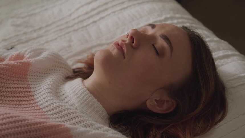Woman with brown hair and a sweater lies down on a bed and opens her eyes