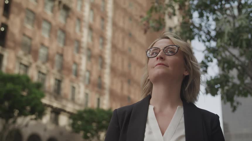 Blonde woman with glasses and a black blazer looks up at tall buildings