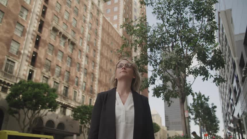Blonde woman with a black blazer jacket and glasses looks up at tall buildings in the street