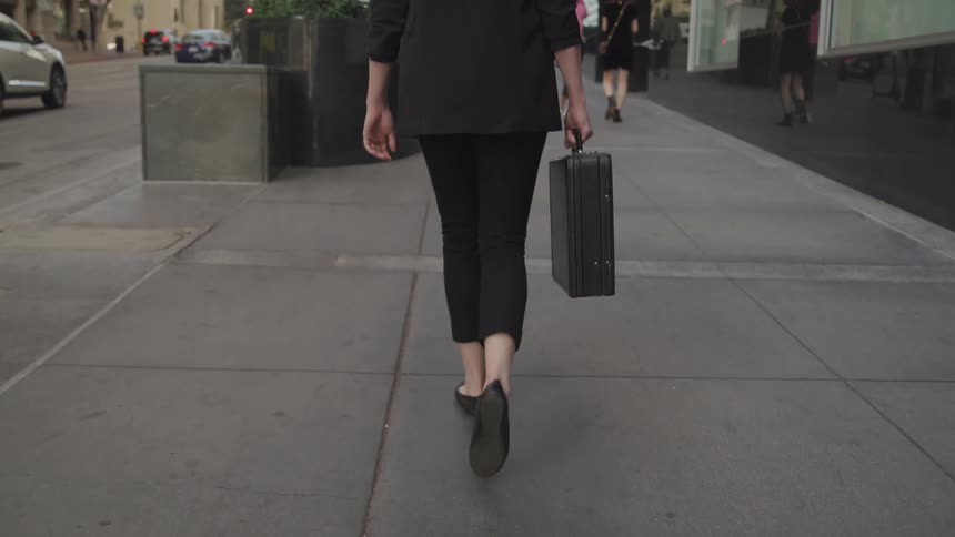 A woman carrying a briefcase walks down the side walk behind other people as cars drive past