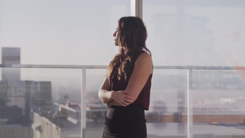 Businesswoman with black wavy hair and a skirt looks out office windows in the office