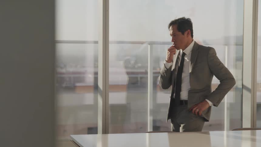 A middle aged man in a suit strokes his chin as he paces an office while lost in thought