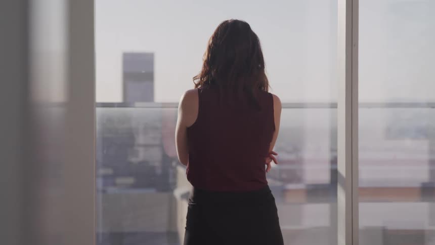 A woman in a black dress stands before a large window looking out over a metropolitan city