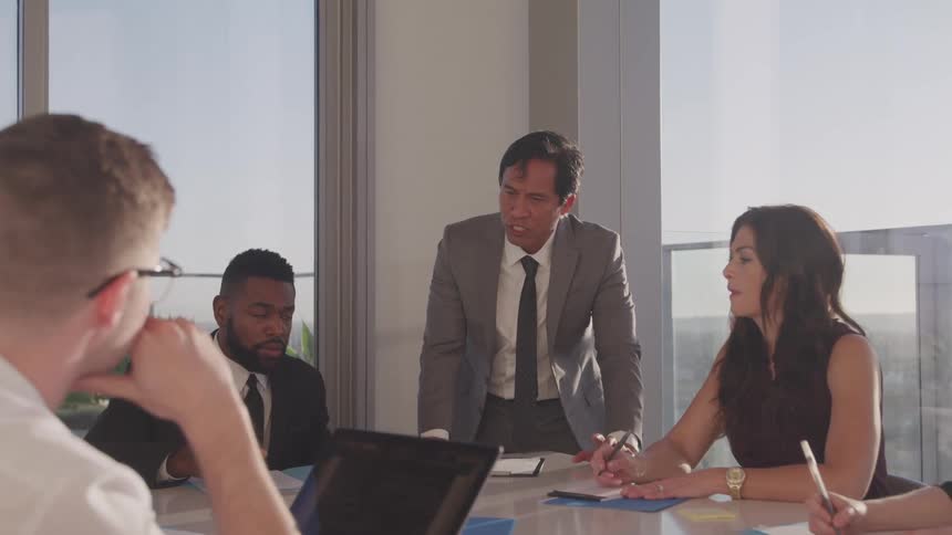A group of employees sit at a table in an office and listen to their boss