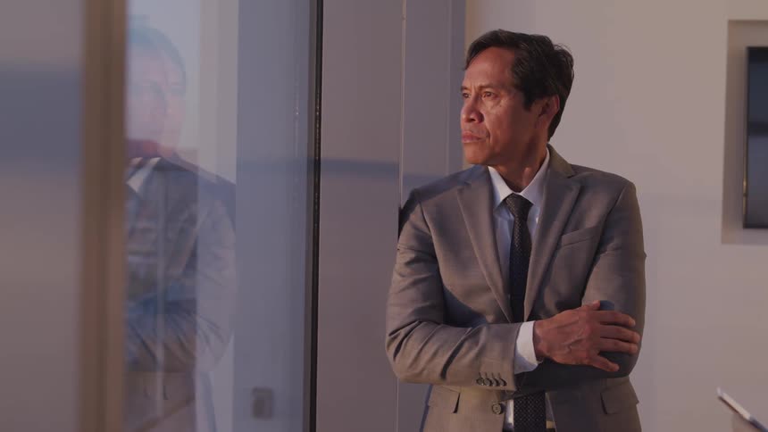 A man in a gray suit stands with his arms crossed as his looks out his office window with views of downtown