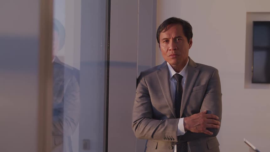 Asian businessman with a grey suit leaning against a wall next to the office windows faces the camera with eye contact