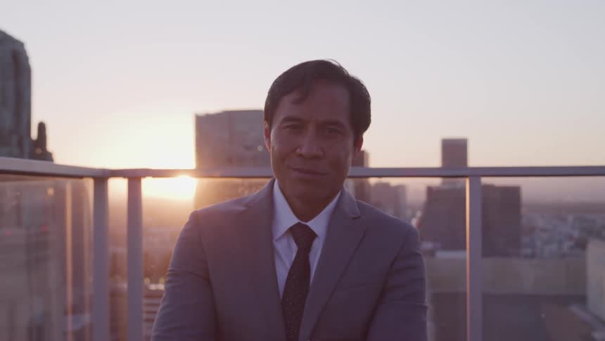 A man with a suit and tie on a building rooftop with buildings and the sky in the background
