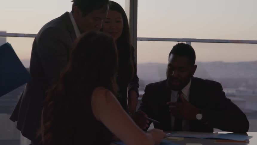 Business people conducting a meeting in a conference room are silhouetted against the sky at dusk