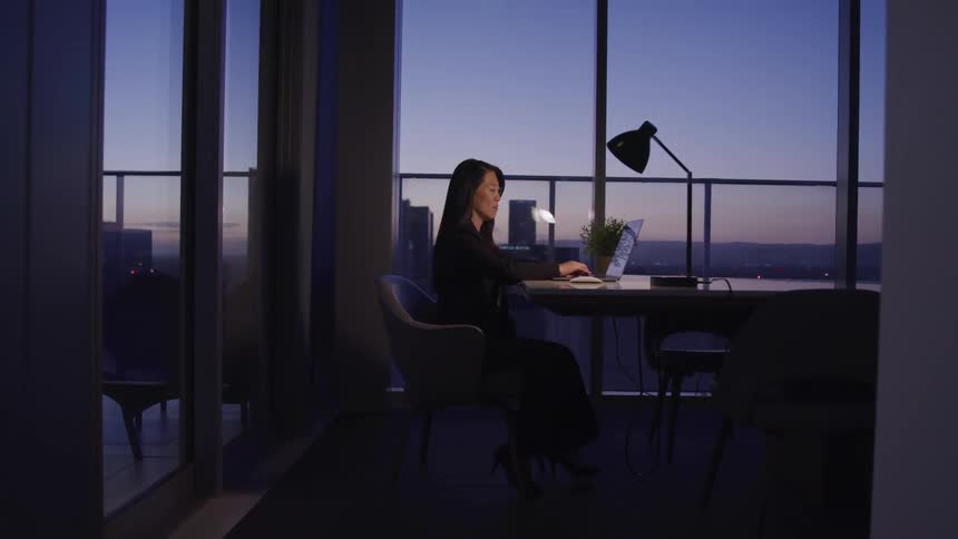 A middle aged asian woman types on a laptop while sitting in an office in a skyscraper at night