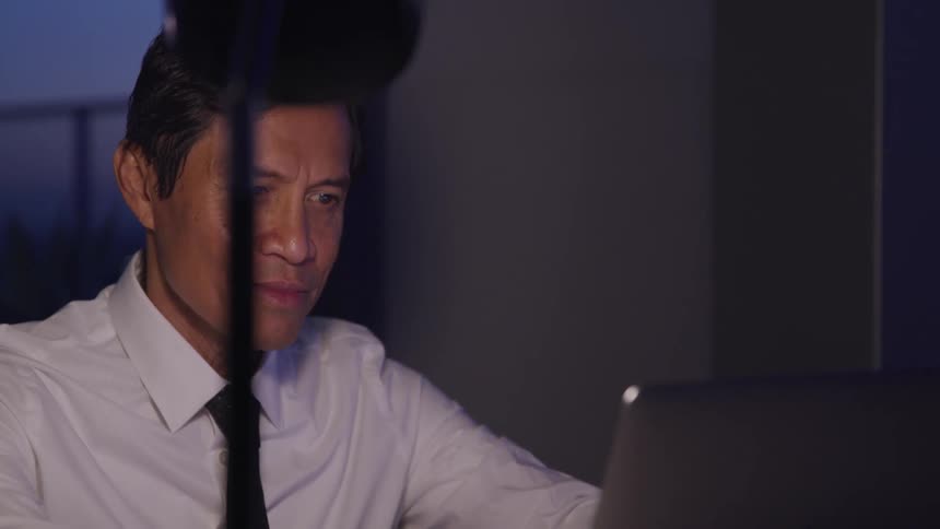 A man looks at a computer inside a room with a lamp and window in it at night