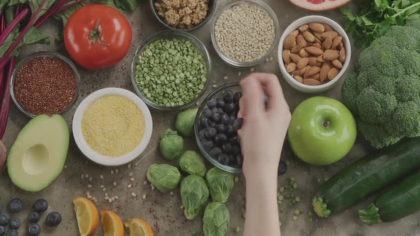 A hand picks up blueberry from a glass bowl sitting next to other fruits and vegetables on a table