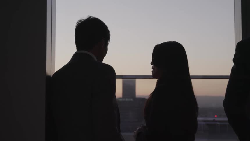Asian businessman and businesswoman standing and looking out the office windows are silhouetted against the sky at dusk