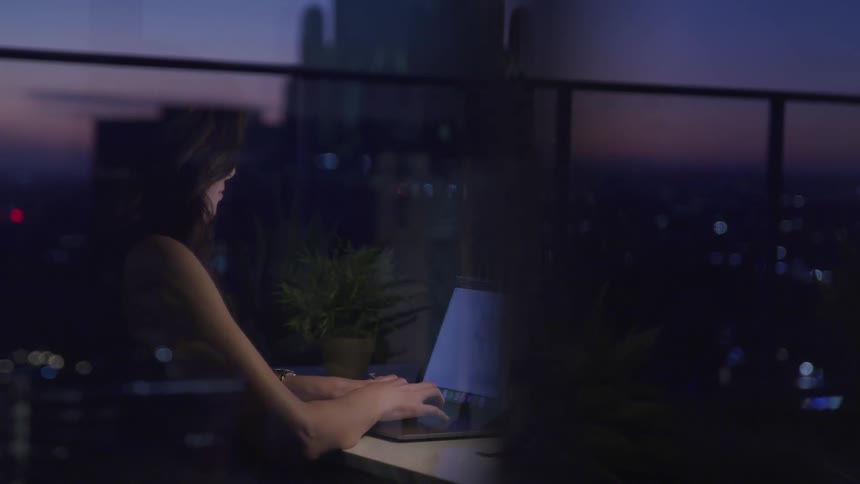 A reflection out a city building window shows a woman sitting at a desk and working on a laptop at dusk