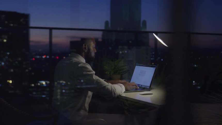 Black businessman with a beard sits at the table and uses a laptop in the office at dusk