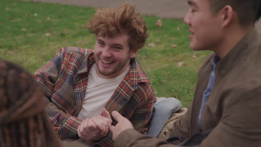 Three friends laugh as they sit on the grass in a park