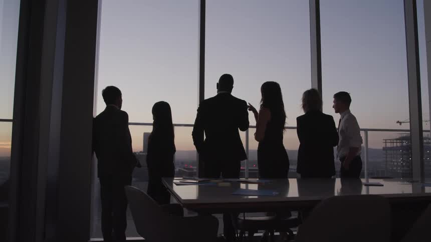 Business people looking out the windows are silhouetted against the sky at dusk in a conference room
