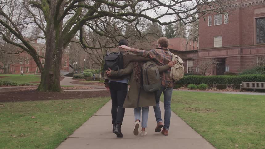 Three friends walking side by side on a campus path put their arms around each other