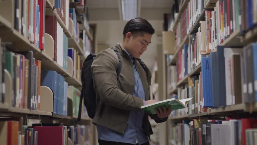 Asian teenager boy stands and reads a book between bookshelves in the public library